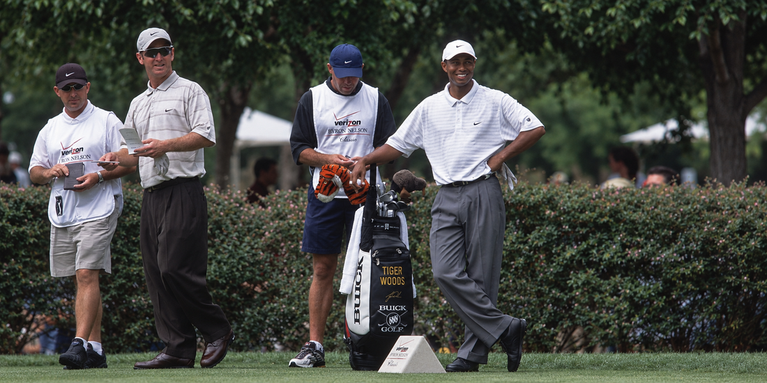 Tiger Woods wearing a white Nike polo and grey trousers stands beside his Buick Golf tour bag at the Verizon Byron Nelson Classic in the early 2000s